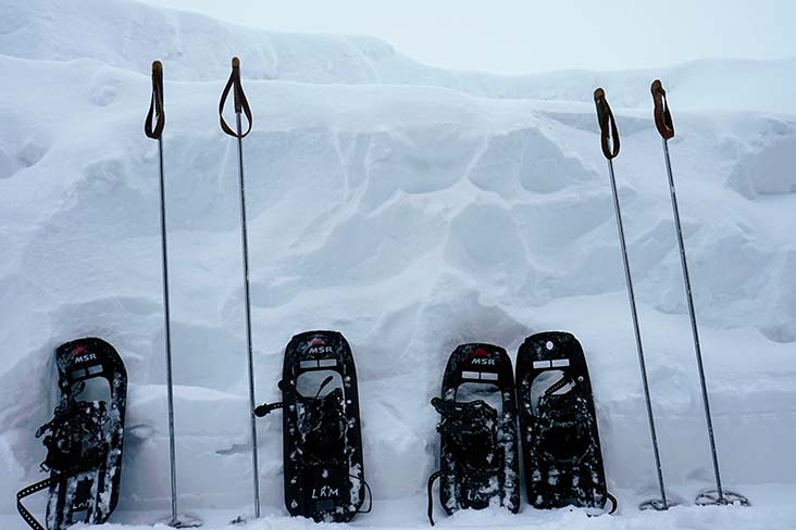 Two pairs of snowshoes and poles leaning against a large snowbank.