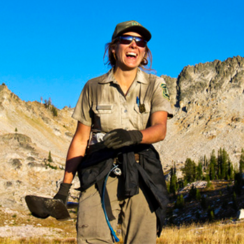 Katy Nelson hiking in a mountainous setting. She’s smiling broadly, and the sky is a brilliant blue behind the mountains.