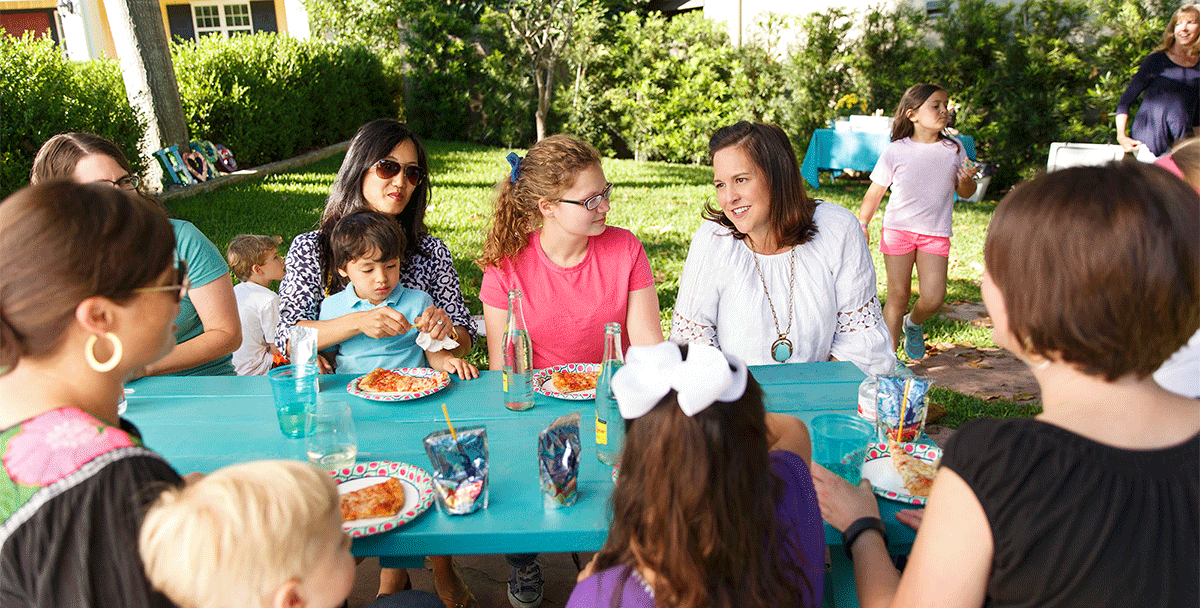 Three women and a toddler enjoy refreshments around a turquoise colored picnic table in a grassy yard