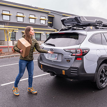 Kimber Cross opening the hatch of her silver Subaru Outback Wilderness, which has a carrier attached to the rooftop.