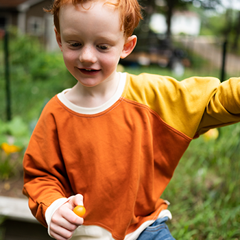 A young child is wearing a Peace House Studio diagonal colorblock sweatshirt. The sweatshirt is made of orange and yellow with white trim.