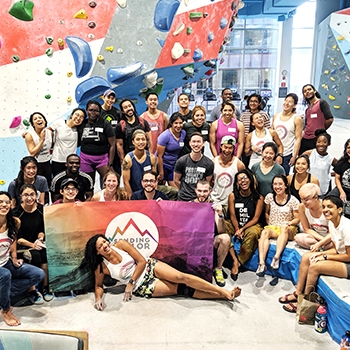 Indira Orozco reclining in front of a Sending in Color banner. She is surrounded by a large group of Sending in Color members, who are smiling. They are inside a gym with rock climbing walls behind them.