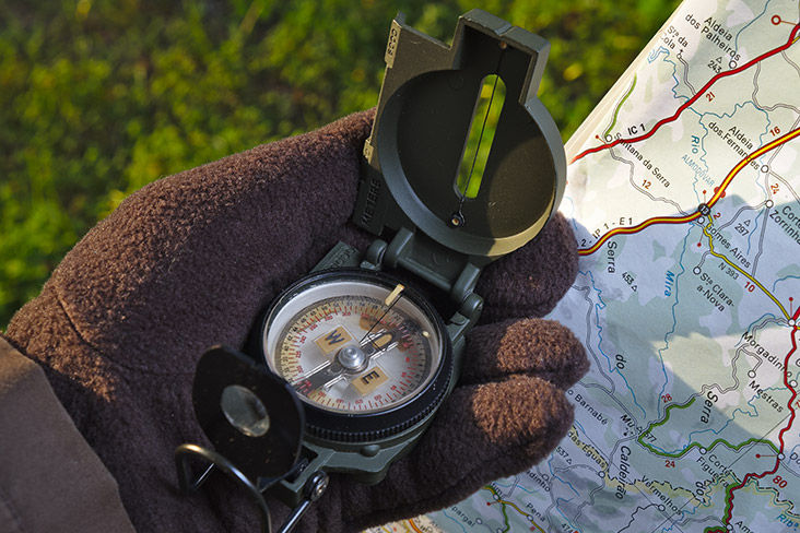 A closeup of a hand that’s wearing a fuzzy, brown glove and is holding a military compass with a paper map behind it.
