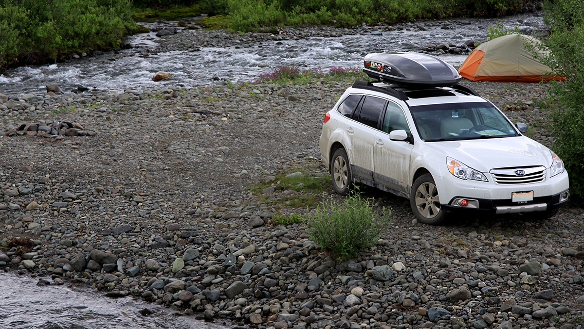 A Subaru Outback with a rootop box parked near the bank of a stream in Alaska