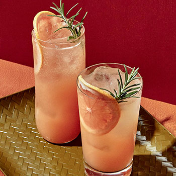 Two tall glasses are filled with a pink-colored beverage, grapefruit slices and ice. Each drink is topped off with a sprig of rosemary.