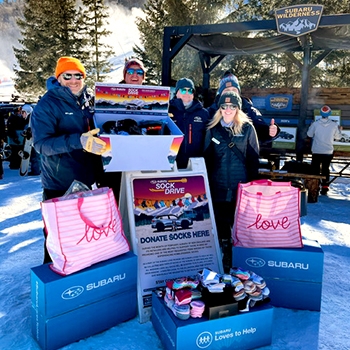 Carrie Cahoon with four other people at a winter event. They're standing in the snow in front of boxes that say Subaru for a sock drive, and some boxes have brand-new socks in or on top of them.