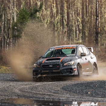 A Subaru with a DirtFish logo across the top of the windshield is turning a corner on a gravel road, kicking up dust and gravel.