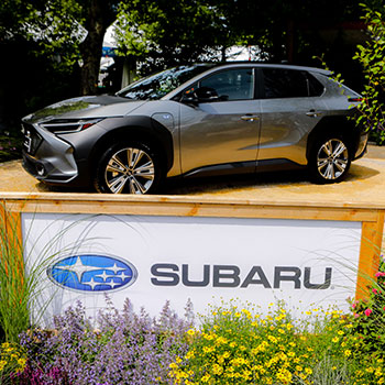 A 2023 Subaru Solterra is parked on a wooden platform behind a bed of wildflowers. On the front of the platform is a sign showing the Subaru logo. 