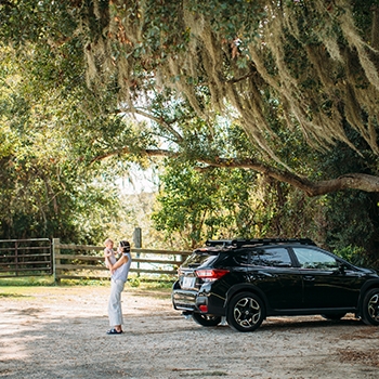 A distance shot of Hailey holding Land in front of their parked black Subaru Outback. Overhead a giant willow tree frames the photo.