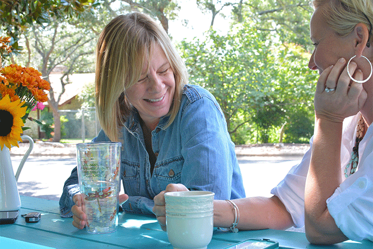 Two women are seated at the turquoise table, smiling.