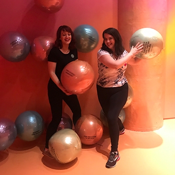 The author and Drive’s art director, both white women, are standing in a peach-pink pastel room holding giant balloons with words on them.