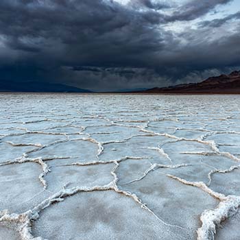 Close-up view of the salt flats of Badwater Basin, California. The salt flats are white with patches of rough, low-lying formations that resemble the roots of trees. Storm clouds loom on the horizon.