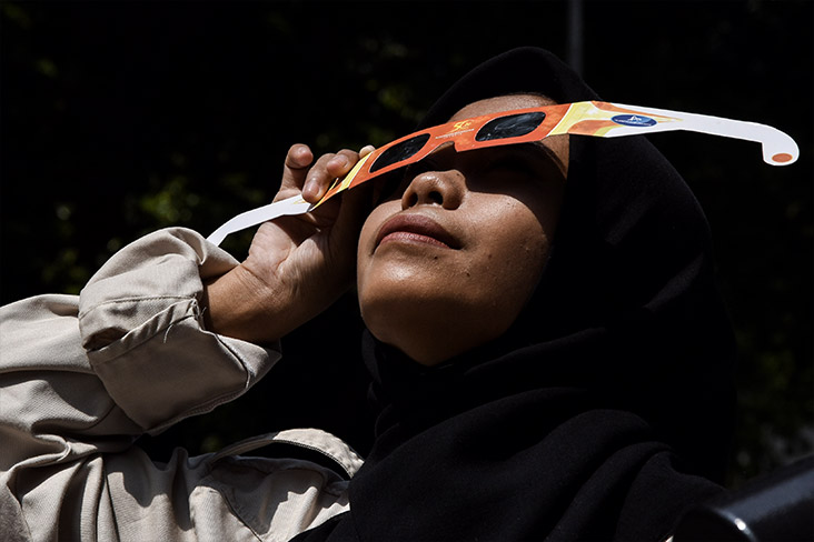 Closeup of a person who is viewing a solar eclipse through a pair of solar eclipse glasses held in one hand covering the eyes.