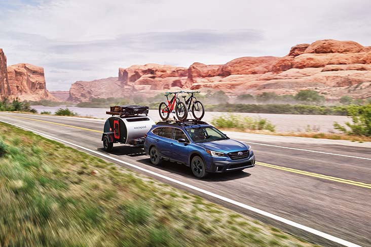An Outback is driving along a canyon road with a Thule Bike Carrier on the roof, which is carrying two bikes. The vehicle is also towing a camping pod hitched on the rear of the vehicle.