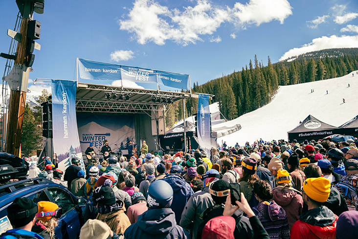 WinterFest attendees are standing in front of a stage listening to music that a four-person band is playing. In the distance, snow covers the mountainside. Groups of evergreen trees are seen beyond the snow and the sky is a clear blue with a few scattered white clouds.