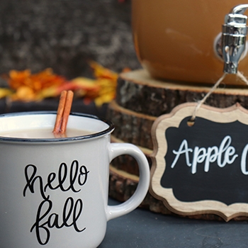 A white ceramic mug that reads “hello fall” and holds apple cider and a cinnamon stick.