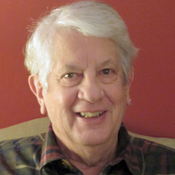 A headshot of Subaru owner Mac Bogert, an older gentleman with white hair, who is wearing a checkered flannel shirt with a collar and is smiling.