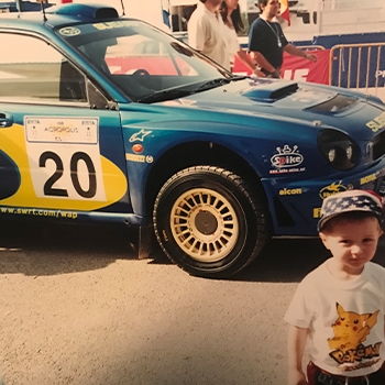 Panagiotis as a young boy wearing a Pokémon T-shirt and standing in front of a World Rally Blue-painted Subaru rally car at the Acropolis Rally in 2001.