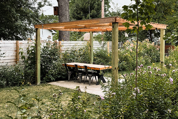 An outdoor dining space in the Kloos’ yard, featuring a concrete patio with a wooden pergola over it. On the patio is a wooden table with six metal chairs. The dining space is surrounded by tall, vibrant native plants.