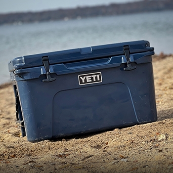 A blue Yeti Tundra 45 Hard Cooler sits on the sand with lake water behind it.