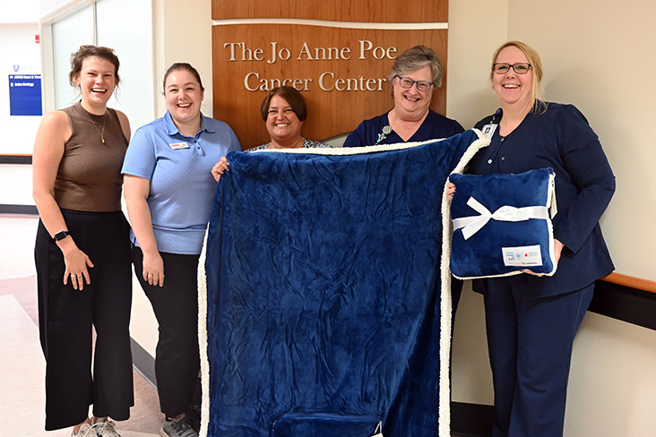 Five women are standing in front of a wall in the Ashe Memorial Hospital in Jefferson, North Carolina, that says, The Jo Anne Poe Cancer Center. They're holding up a large velvety blue blanket and pillow and are smiling.