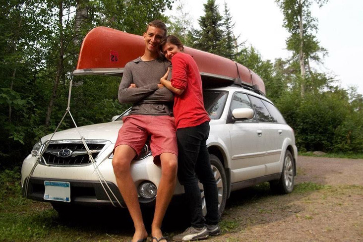 Karl and Katie Kloos with their 2009 Subaru Outback. Karl is leaning on the bumper of the vehicle, and Katie is next to Karl embracing him. They are on a gravel road in a forested area, and a canoe is on the roof top of the Outback.