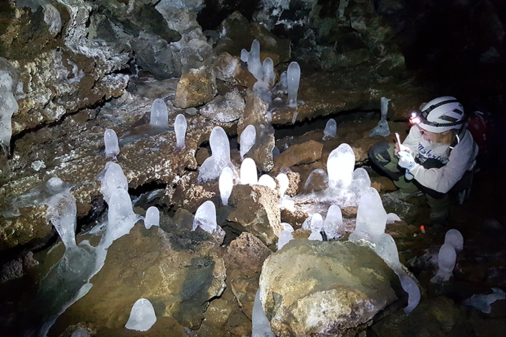 Ice formations that look like bowling pins on the cave floor. On the far right, a person wearing a helmet is squatting in front of a formation and taking a picture.