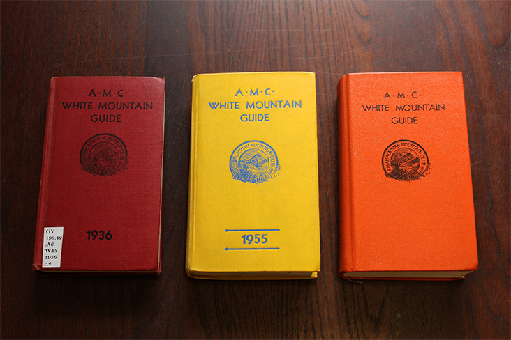 Three hardcover examples of the AMC White Mountain Guide are spread out on a table. From left to right the book colors are red, yellow and orange, and the earliest version dates back to 1936.