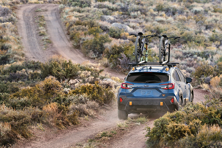 A Subaru Crosstrek Wilderness with two bikes mounted on the rooftop. It is driving on a dirt road with vegetation on each side of the road.