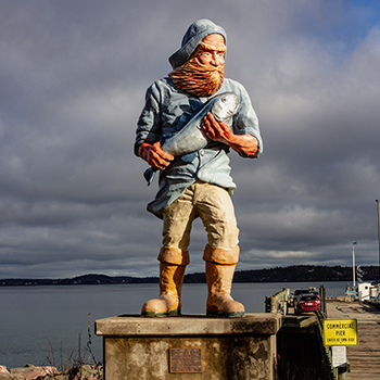 A statue of a fisherman who is wearing knee-high boots, khaki pants and a blue shirt and hat. His long red beard is sculpted to look as if it’s blowing in the wind. Behind the statue, the sky is cloudy and water can be seen in the distance.