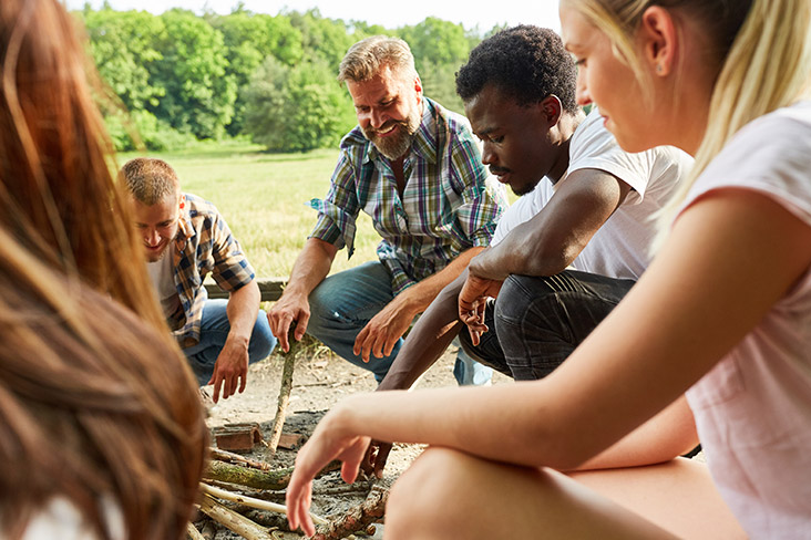 A small group of men and women are in a circle, smiling. They are in a clearing near the woods and are building a fire with sticks. Some are wearing white T-shirts and others are wearing flannel shirts that have the sleeves rolled up. 