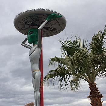 A larger-than-life-sized statue of an alien figure thatʼs holding up a UFO, which was taken outside of the Alien Fresh Jerky store in Baker, California.
