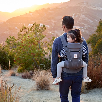 A father carries his daughter in the baby carrier on his back, both facing forward looking at a sunset over a mountainside
