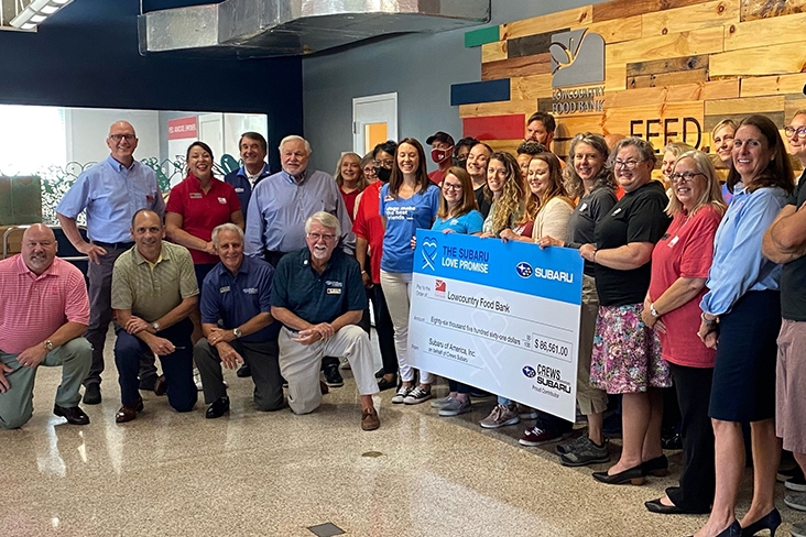 The Crews Subaru team presenting an oversized check to Lowcountry Food Bank. There are about 25 people standing or kneeling around the check donation. The check is for $86,561.