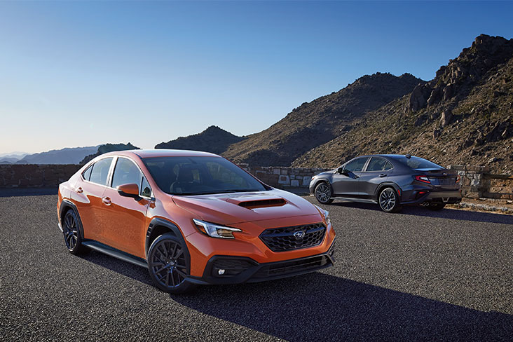 In the foreground, an orange 2022 Subaru WRX Limited is parked on asphalt. In the background, a gray 2022 Subaru WRX GT is parked at the edge of the asphalt near ragged mountains.