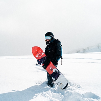 A snowboarder walking through fresh snowfall, wearing goggles and a black winter coat, holding a snowboard and looking back at the camera.