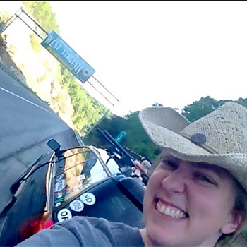 Amanda Isaacs, wearing a cowboy hat, stands in front of her Subaru Outback taking a selfie. Beyond the Outback is the sign welcoming visitors to West Virginia. 