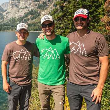 Sean Stowers, Ben Kieffner and Brian Stowers are standing arm in arm in a wilderness setting. A body of water is behind them, and they are surrounded by greenery.