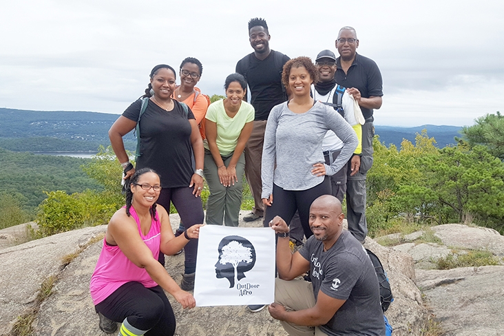 Kasim Carter with a group of eight posing on a boulder with an Outdoor Afro banner displayed. In the distance are deciduous trees and a body of water.