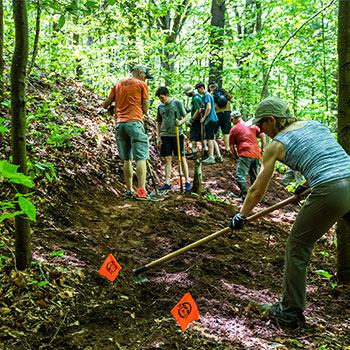 A group of New England Mountain Bike Association members are hoeing a muddy trail in a wooded area.