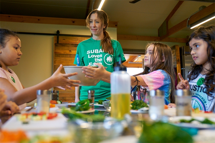 At Apple Seeds educational farm, Danielle Dotson, who is wearing a green Subaru Loves the Earth T-shirt, stands next to a table. Three adolescent girls are seated at the table and are working on an activity together.