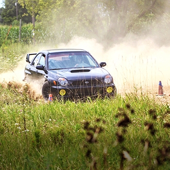 Derric Throne, co-driver for the Art in Motion Rally Team, driving his Subaru through one of the courses at FL4TFEST. There is a lot of dust behind the vehicle and tall grass in the foreground.  