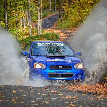A blue vehicle is driving along a forest road and spraying up dust and gravel into the air. Across the windshield it says, Team O’Neil Rally School. There are many trees on both sides of the road behind the vehicle.
