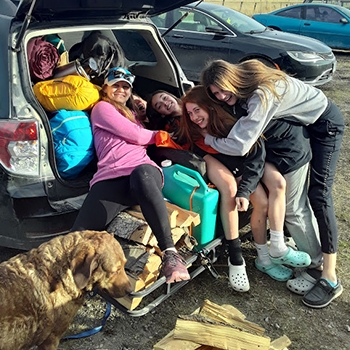 Lynn Rose Pine, wearing a long-sleeved pink shirt and a cap with sunglasses over it, plus four female teens, are tumbled in the back of a packed Subaru Forester, smiling and hugging each other. A medium-size dog stands nearby.