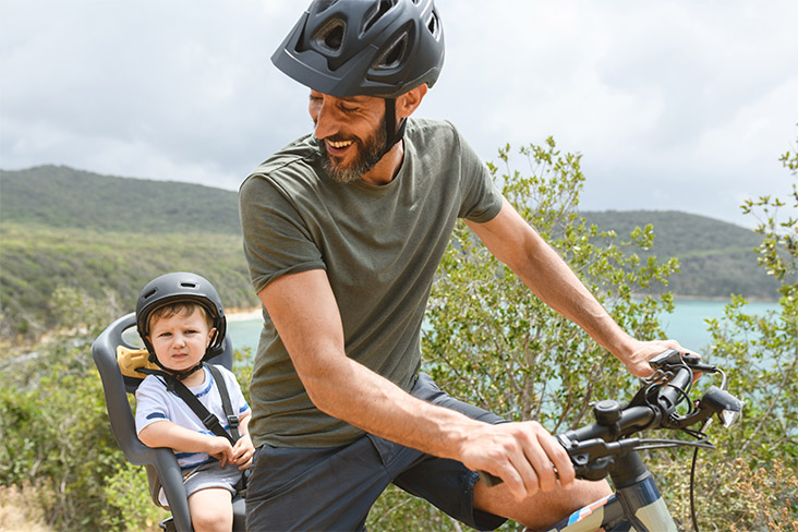 A man is on an e-bike with a toddler harnessed in a seat behind him. He is looking back at the child, smiling. Both are wearing helmets. They're in a country setting with a lake and green hills in the distance.