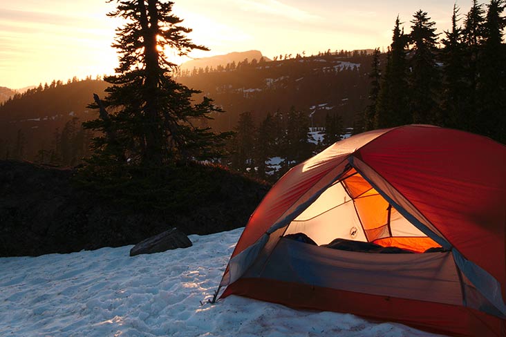 Tent pitched in snow with evergreens and sun setting in background.