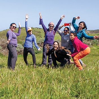 A group of seven women, dressed in colorful outfits for outdoor adventure, strike goofy poses for the camera outside on grass in the sunshine.