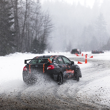 A Subaru is driving on a winding DirtFish course in the snow. In the distance, some orange cones have been knocked over. The vehicle’s wheels are leaving a trail of snow in the air behind it.
