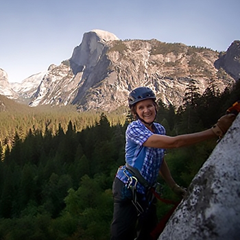 Dierdre Wolownick, wearing a helmet and smiling as she climbs up a rockface with a mountain and trees in the background.