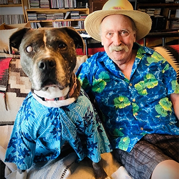 Mr. Piggums is on the left, wearing a Hawaiian shirt, and Woody Konopelli is on the right, also wearing a Hawaiian shirt. Mr. Piggums is looking eagerly at the camera, and Konopelli is smiling next to him. A bookshelf filled with books can be seen in the background. 